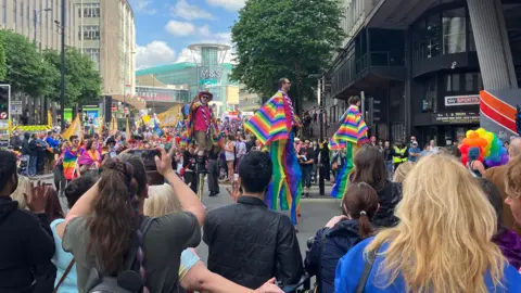 Birmingham Pride celebrations in the city centre. Crowds gather and rainbow flags fly. Street performers on stilts walk past. 