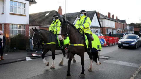 Getty Images Two female police officers ride down a residential street in Cheltenham on bay horses. They are wearing uniform, including hi-vis yellow coats and surgical masks. Two cars are driving behind them.