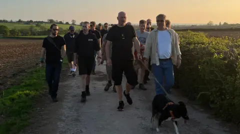 A large group of men, one of which is walking a dog, walk through a field on a path at sunset. 