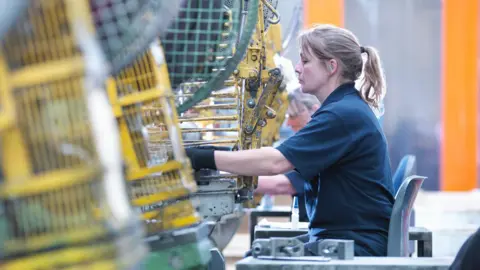 Getty Images A woman wearing a blue polo t shirt and black gloves operating a punch machine in an engineering factory