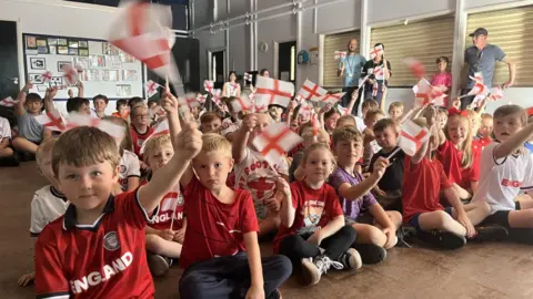 BBC Primary school children sit on the floor with their legs crossed waving England flags. All are dressed in red and white. 