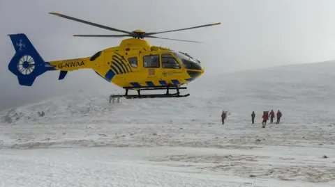 Keswick Mountain Rescue Team A team of Keswick Mountain Rescue volunteers, who are all wearing red waterproofs and large rucksacks, stand on top of the summit of Blencathra. There is a yellow helicopter hovering above them.