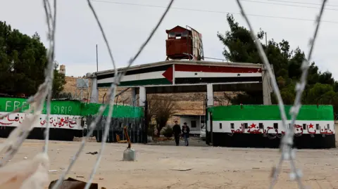 View of a prison through barbed wire, with two people walking and walls painted with 'Free Syria' and the flag colours