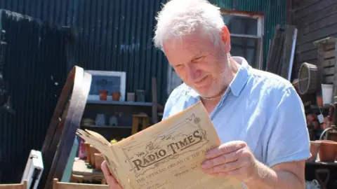Caty Mills Steve Hopkins holds open the 101-year-old copy of the Radio Times in his hands as he reads it in the yard of his vintage business. He is surrounded by plant pots, mirrors and other items. He has white hair and a blue short-sleeved shirt.