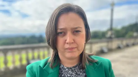 Deborah Erskine pictured outside Stormont, with a stone wall behind her. She has shoulder length brown hair and is wearing a green jacket and black and white floral top. She is looking directly into the camera. 