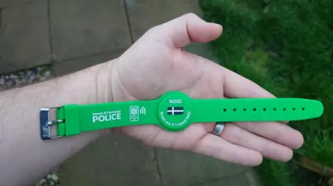 A hand laying flat. There is a wristband lying on the palm of the hand. It is green, has a Cornish flag in the centre and says "NDID" and "Scan me if I need help" in the centre. On the left strap it says "Devon and Cornwall Police" with a picture of a phone next to it. 