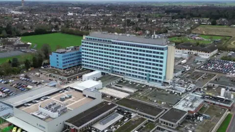 An aerial shot of a hospital estate, which is dominated by a large 10-storey building with blue cladding and rows of black windows. It is surrounded by smaller one and two-storey buildings and car parks. Green playing fields and houses can be seen in the background.