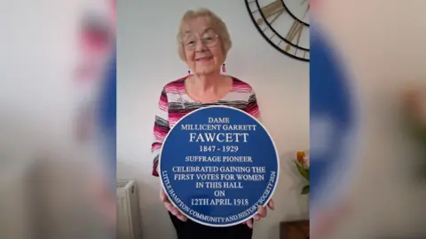 Christina Merry The image shows a woman holding up a blue plaque commemorating Suffragette Dame Millicent Garrett Fawcet.