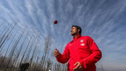 Abid Bhat Wearing a bright red jacket, Parvez Rasool stands in an open field as he flicks a red ball up in the air. The sky behind him is blue and full of long clouds