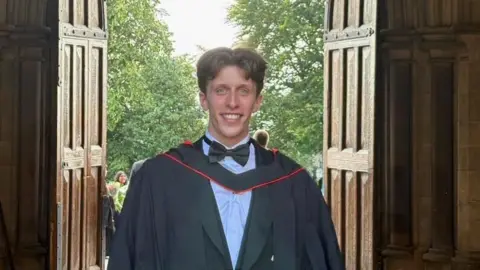 Sam Gurling Sam wearing a black graduation gown with red trim and a bow tie stands in a large stone doorway, with sunlight shining through and green trees visible outside