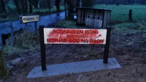 Red spray paint obscuring bilingual text on a road sign in the Fermanagh and Omagh council area. The road, a bus stop and a field can be seen in the background.