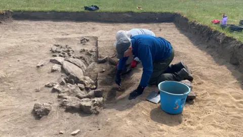 Two people are on their hands and knees in a shallow archaeology trench. In front of them are some stones and they are scraping the dirt from around them. There is a blue bucket is next to them 