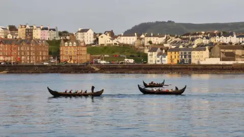 Jimmy Lee Three brown Viking longboats sail next to each other in Peel Bay. You can see rows of houses and green hills in the distance.