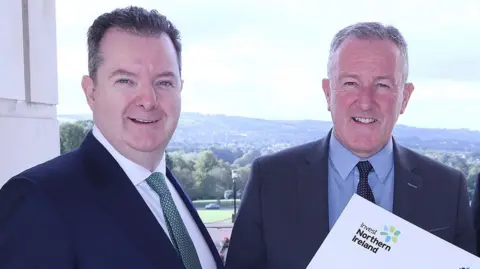 Kieran Donoghue and Conor Murphy standing side by side on a balcony at Stormont, you can see green hills in the background.
Both are wearing suits and ties.
The edge of an Invest Northern Ireland logo can be seen.