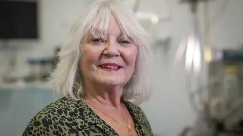 BBC Doreen Adams, una mujer con cabello gris hasta los hombros y raya en medio, sonríe y viste una blusa y un collar verde y negro, con equipo médico al fondo.