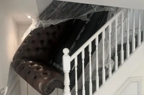 Luke Ansell A chair, still partly in wrapping, is lodged in a stairwell between the sloped roof, wall, and banister.