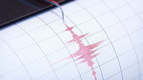The photo shows a needle which is recording a series of jagged, up-and-down squiggly lines on a moving roll of paper in red ink. 