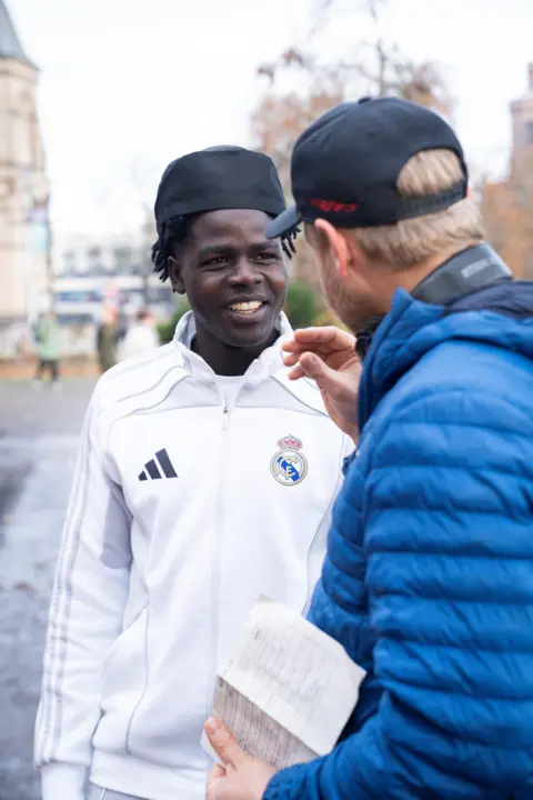 Compass Collective Two men are talking next to the riverside in Inverness. In the foreground we can see a man in a blue jack although we can't see his face because he is facing away from the camera. He is talking to a young black man who is smiling. He is wearing a black hat and a white football tracksuit.
