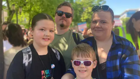 Two children and their parents.
The girl has a black tshirt and bag on. She has black hair in a middle parting.
The boy has blond hair and purple glasses on.
The woman has a blue shirt on and sunglasses on her head.
The man has blond hair and a green shirt on with black sunglasses.