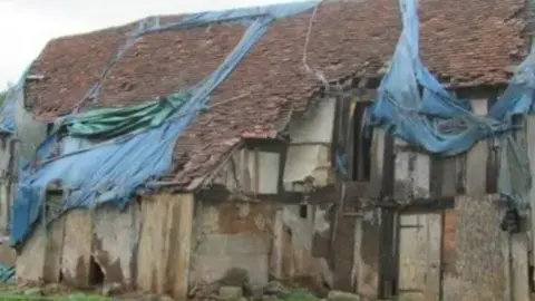 A delapidated looking building - it looks to be a timber building with some boardings and some blue protected sheeting on the roof which has come away and is dangling down the front of the building. Red roof tiles can be seen 