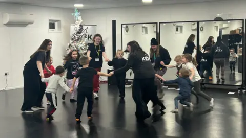 A group of children holding hands in a circle in a community centre dancing