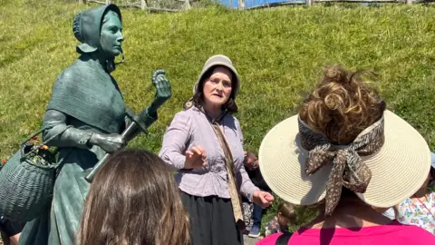 A woman is dressed in Victorian clothing and giving a talk to crowds while stood in front of a statue of Mary Anning.