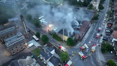A large plume of smoke is seen from above. It is pouring from the roof of a large supermarket. 