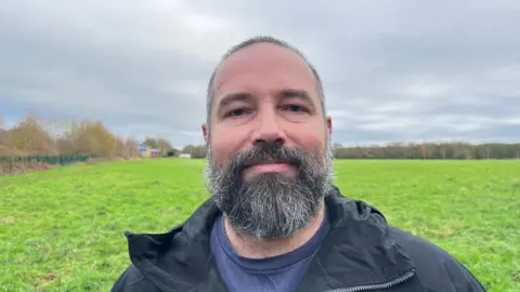 Steve Bryant looking down the camera, wearing a black raincoat with a dark blue top underneath. Behind him is the green field that will be built on. In the far distance you can see the tree line. The sky is grey and cloudy.