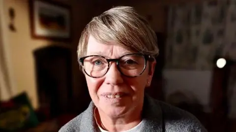 Amanda McKay, in her late 50s, with glasses and a grey top, with a dark background  