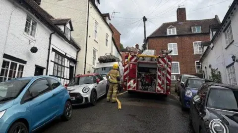 A fire engine is parked on a steep, narrow street, with two sets of cars either side of it. There is also a firefighter in full gear and a hose on the floor. There are terraced cottages on either side of the street.