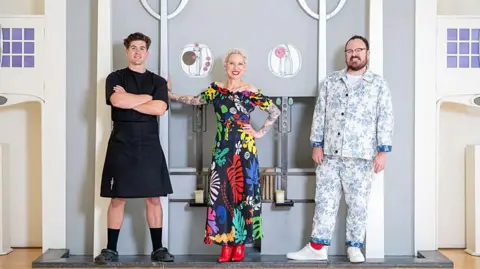Danny Campbell, Anna Campbell-Jones and Banjo Beale stand in a line in front of a beautiful fireplace inside the House for an Art Lover, where the winner was announced. Danny wears a black t-shirt, black kilt and Crocs, Anna, with her bright white cropped hair, wears a bold floral dress and red boots and Banjo rocks a blue and white china pattern-style suit with white trainers.