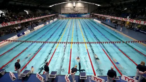 PA Media An Olympic-sized swimming pool has competitive swimmers taking part in a race in lanes with adjudicators watching poolside from the ends of their lanes