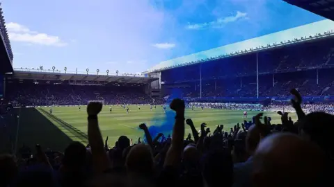 Getty Images View from inside one of the ends at Goodison Park. Arms are raised in silhouettes from among the crowd, and there is blue smoke from a pyrotechnic. There are players on the pitch.