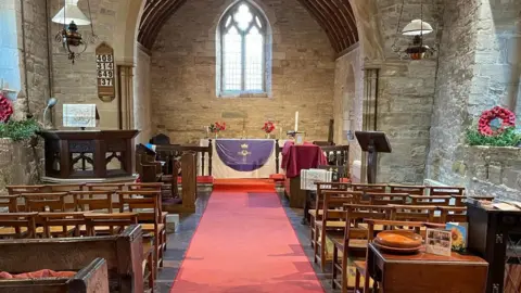 Rev Ian Perry The inside of a church which has a red carpet and wooden pews on either side. The alter at the front has a white and purple cloth on it and there are candles. 