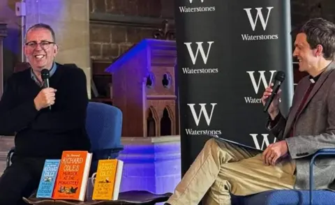 Diocese of Peterborough Richard Coles with short dark hair and glasses, smiling while holding a microphone and wearing a black sweater. The Reverend Tom Houston, with short dark hair and a grey jacket, sits opposite. Books by Mr Coles are on display in the foreground, and a Waterstones logo is visible behind