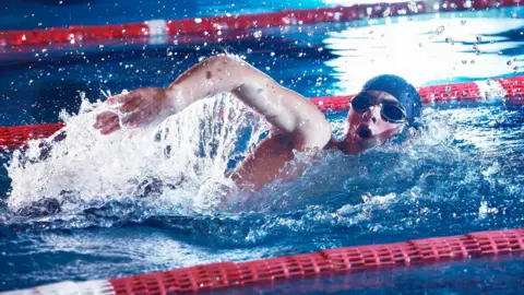 A stock image of a man wearing goggles and a swimming cap swimming in a pool.