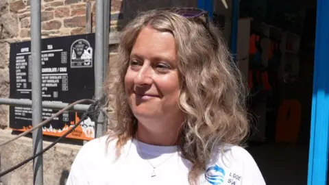 Lauren Pattison from Looe Beach Hire looking away from the camera and smiling. She is wearing a white branded shirt and is stood in the sunshine. 