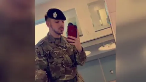 Contributed Selfie of Kyle Clifford wearing his army uniform and beret in a bathroom. He is holding a red phone and has a dark, close-cropped beard and moustache.