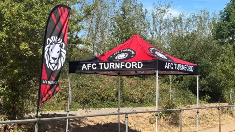 AFC Turnford A flag and gazebo showing AFC Turnford, by a football pitch. It is made out of red and black colours. There are trees behind it and an area of grass, that is dried up. 