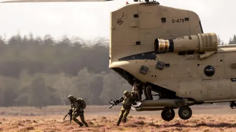 Dutch soldiers of the 11th Airmobile Brigade leaving a Boeing CH-47F Chinook helicopter. The large beige coloured aircraft has yet to fully touchdown, and it's rear wheels are still inches from the ground as two soldiers leap out of the vehicle. 