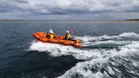 Seahouses RNLI An inshore lifeboat containing three people all wearing hi-vis gear and lifebelts is speeding across the sea with a lot of wash behind it. In the distance you can see a small lighthouse 