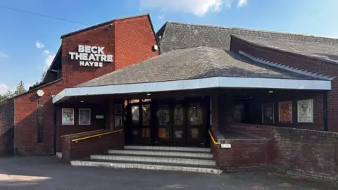 Philip James Lynch/LDRS A 1970s brick theatre building with a sloping tiled roof. There are several steps leading to the door, and yellow handrails. A sign reads: "Beck Theatre Hayes".