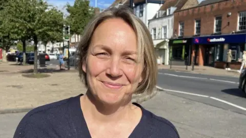 Simon Dedman/BBC Kerry Gilroy is looking at the camera on a sunny day. She has a short blonde bob and is wearing a dark blue top. Shops can be seen in the background as well as people walking on a street and a car on the road