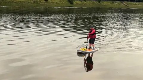 Woman paddleboarding toward the camera. She is standing up holding a paddle and wearing a bright pink waterproof coat, black trousers, a black cap and black lifejacket.