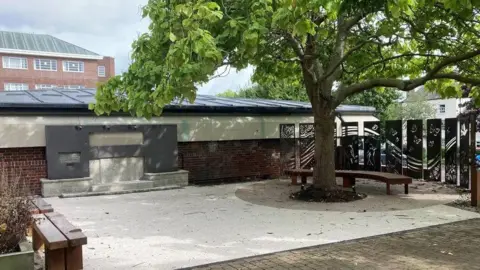 A memorial square with benches and a tree in the middle of a paved area. There are panels on the right hand side covered in designs.