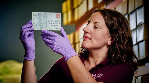 English Heritage A woman wearing purple gloves holding a postcard. The handwritten back of the postcard is visible, which has a very old stamp and illegible handwriting.