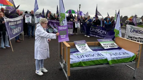 Multiple people holding purple and green unison flags and a hospital bed covered in unison signs sits outside Stormont and is being held on my a protestor. 