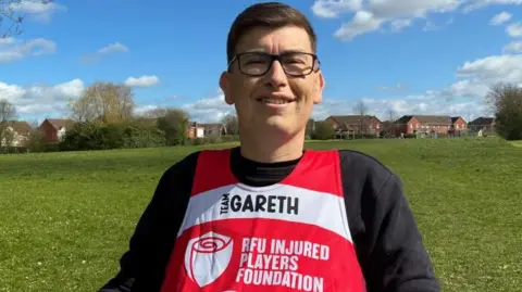 Gareth Rees sits in his adapted wheelchair with a park and houses behind him on a sunny day with a blue sky and white fluffy clouds.