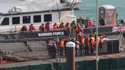 People wearing orange life jackets standing on a white boat. There are people standing on a pier. 