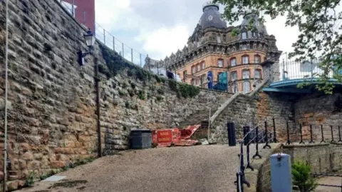 North Yorkshire Council A sloping pathway bordered by stone walls. The pathway is paved with a textured surface, and along the right side runs a black metal railing. At the top of the incline, there is an orange barrier with visible text that reads “ROAD CLOSED”.